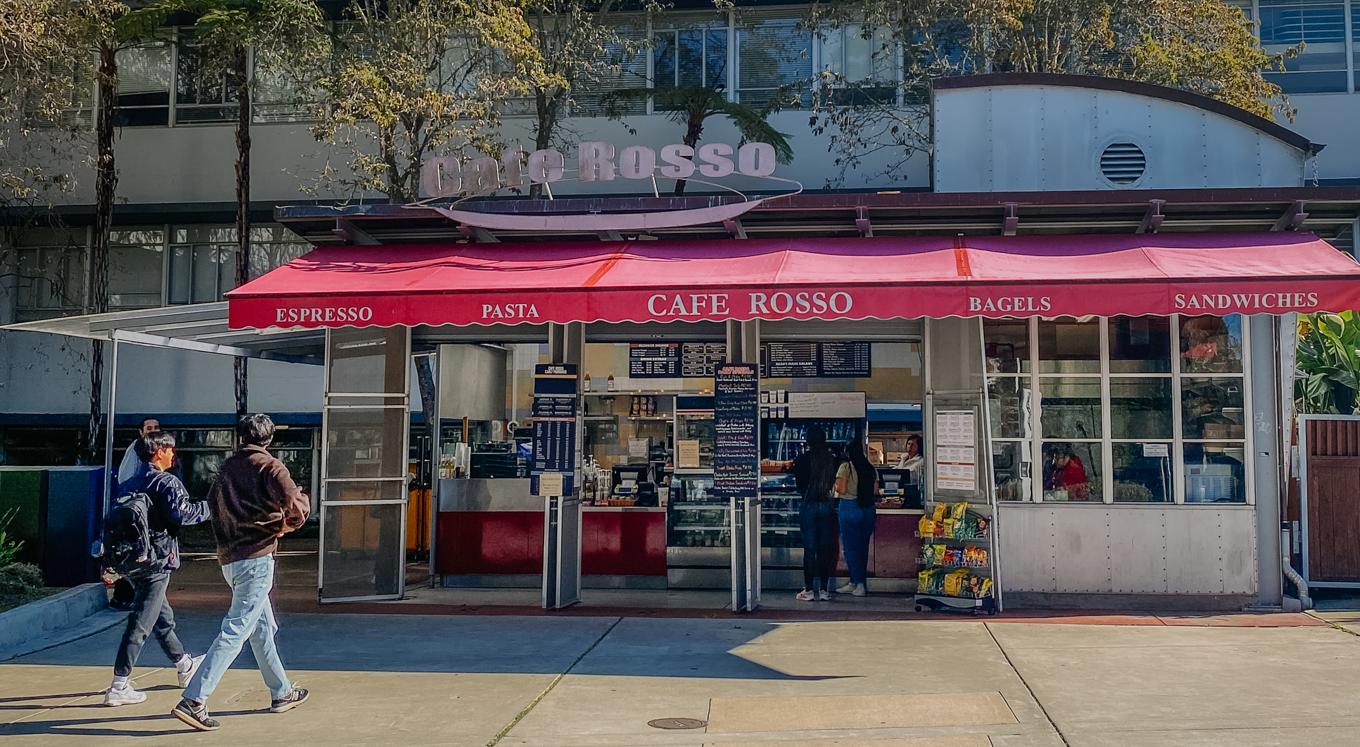 Two students walking towards Cafe Rosso, the sun shining on its red awning.